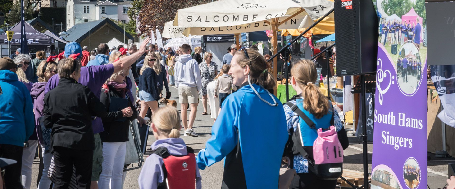 People at an outdoor event with market stalls, a sign, and people wearing various clothing.