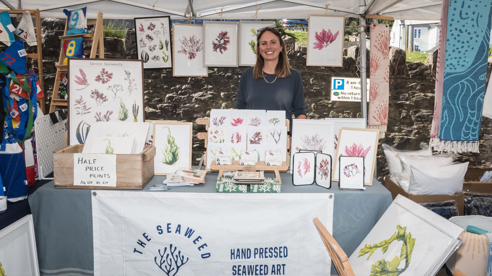 Woman at an art market stall selling framed pressed flower art. Blue and white banner.