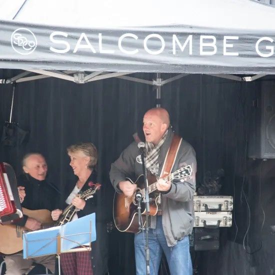 Band performing outdoors under a tent: man singing and playing guitar; others play instruments, Salcombe sign.