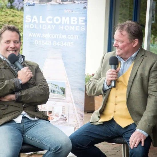 Two men seated, each holding a microphone, in front of a banner for Salcombe Holiday Homes. They are smiling.