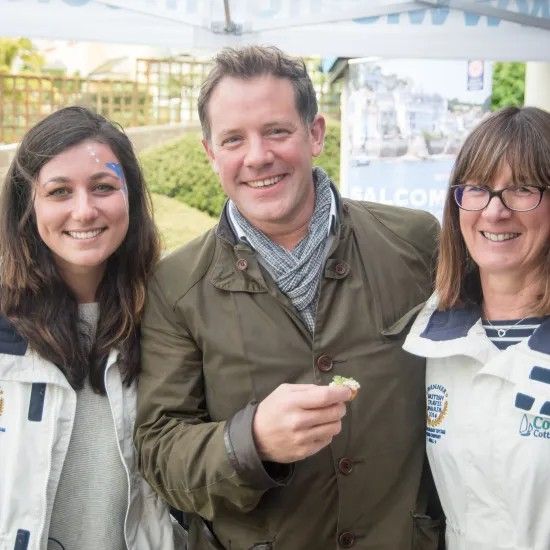 Three smiling people posing outdoors; man holding food, women wearing jackets.