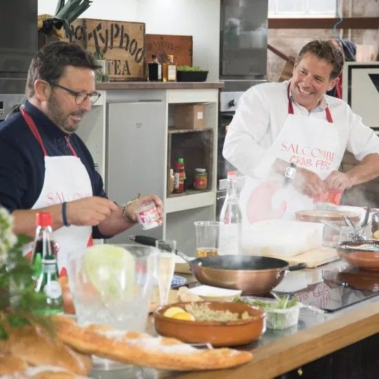 Two chefs in a kitchen; one adds ingredients, the other chops. They wear aprons and smile.