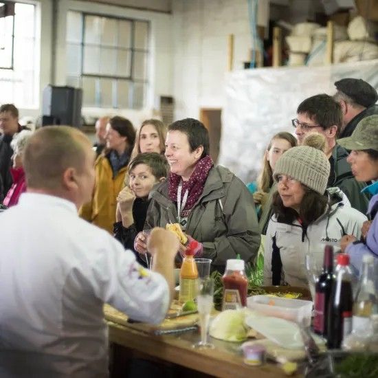 Chef speaking to a group of people at a counter; food and bottles visible.