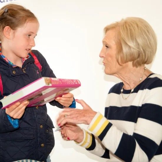 Girl handing a cookbook to a woman with blonde hair, who is signing it. White wall backdrop.