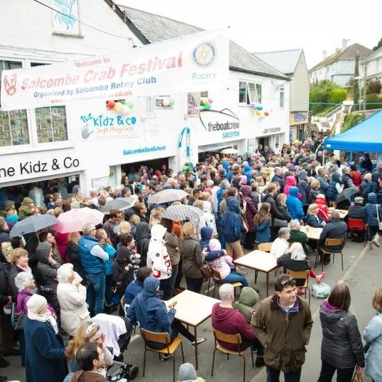 Crowd at Salcombe Crab Festival, outside shops and tables in street. Some umbrellas, cloudy day.