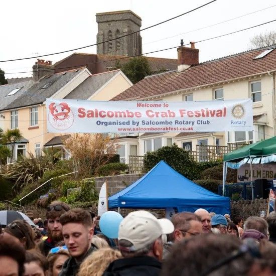 Crowd at Salcombe Crab Festival, banner over street, blue tent, beige buildings, church tower in background.