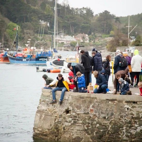 People fishing from a stone pier in a harbor, boats in the background.