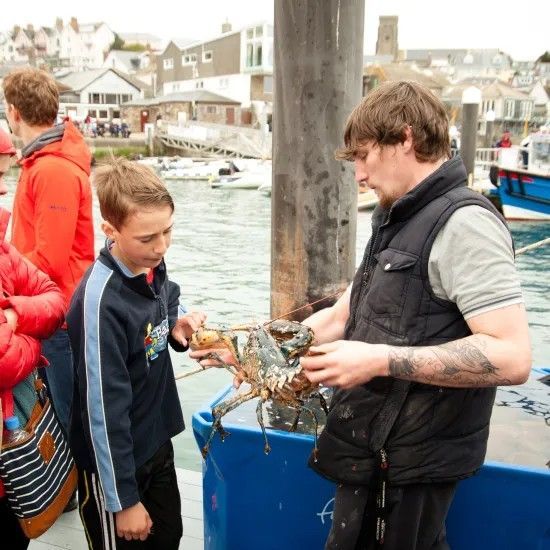 Man showing a lobster to a boy at a harbor, buildings and boats in the background.