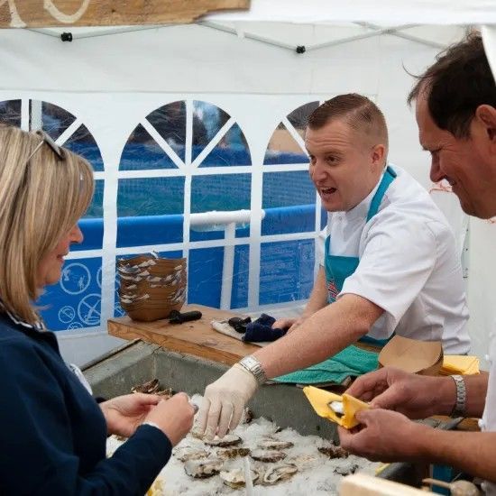 Oyster vendor serving a customer at an outdoor market, with oysters on ice, blue and white tent.