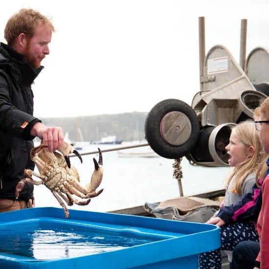 Man holding a crab, showing it to excited children near a blue water tank on a boat.