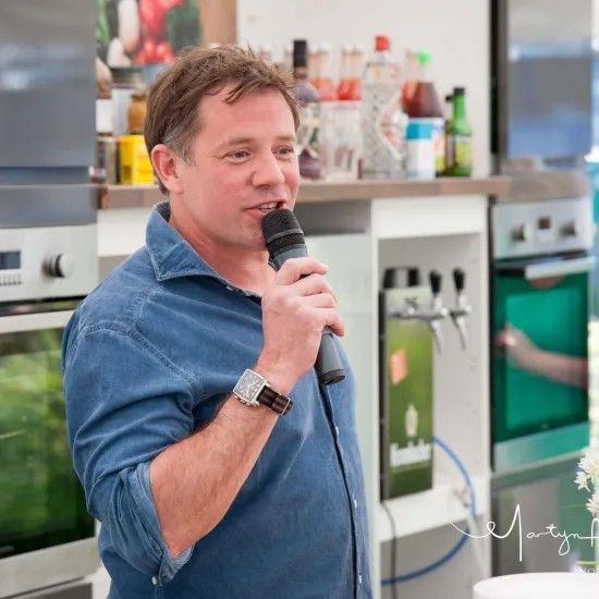 Man in blue shirt speaks into a microphone in a kitchen setting.