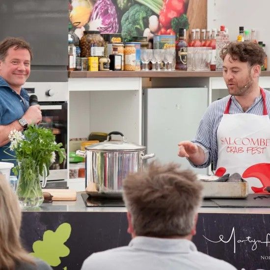 Two men at a cooking demonstration. One with a microphone, the other cooking with a large pot.