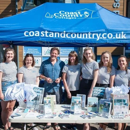 People at a Coast and Country Cottages promotional stand. Blue tent, brochures, and staff in striped shirts.