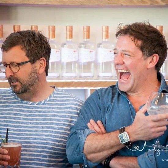 Two men at bar, one laughing with drink, other with glasses holding a cocktail. Bottles in background.