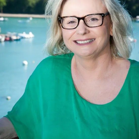 Woman with glasses smiles; green shirt, water and boats in background.