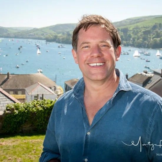 Man smiles, standing outdoors overlooking a bay with boats. He wears a blue shirt.