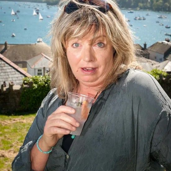 Woman holding a drink outdoors, overlooking a coastal scene with boats. She is wearing a grey shirt.
