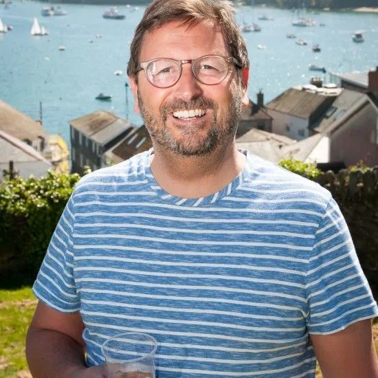 Man with glasses and striped shirt smiles, holding a drink, with a seaside view in the background.