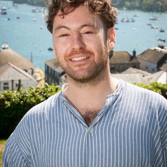 Man with curly hair smiling, wearing a striped blue shirt, background includes a coastal town.