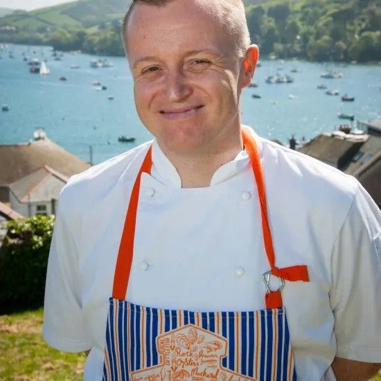 Chef smiling outdoors by water; wears white coat and blue striped apron with orange straps.
