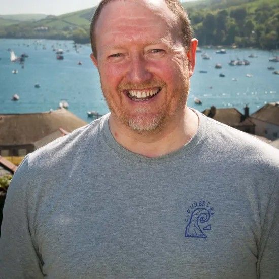 Man smiles at camera, wearing a gray t-shirt with a blue logo, standing outdoors with a bay and boats in the background.