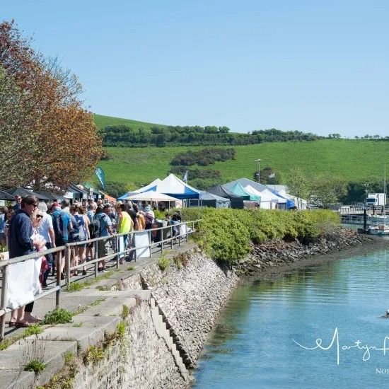 People walking along a waterfront, shops and tents in the background, green hills and blue sky.