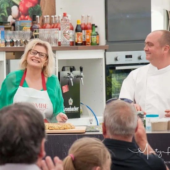 Woman in glasses and man in chef's whites at a cooking demonstration. Smiling, apron, food on counter, crowd in front.