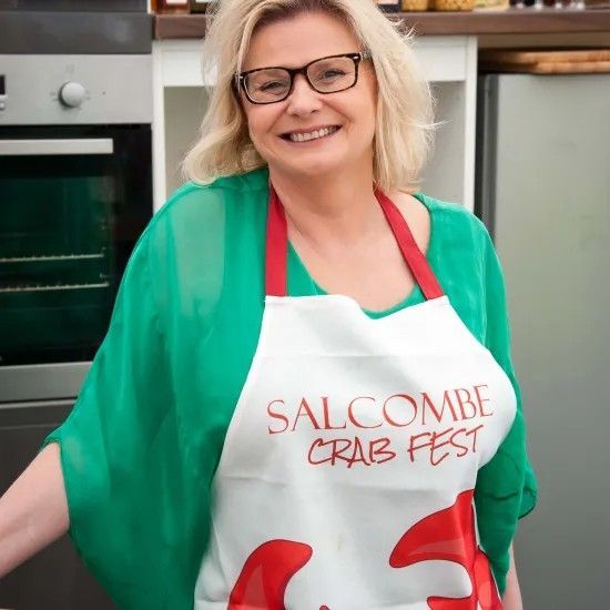 Woman in green top and apron smiles in a kitchen, promoting 