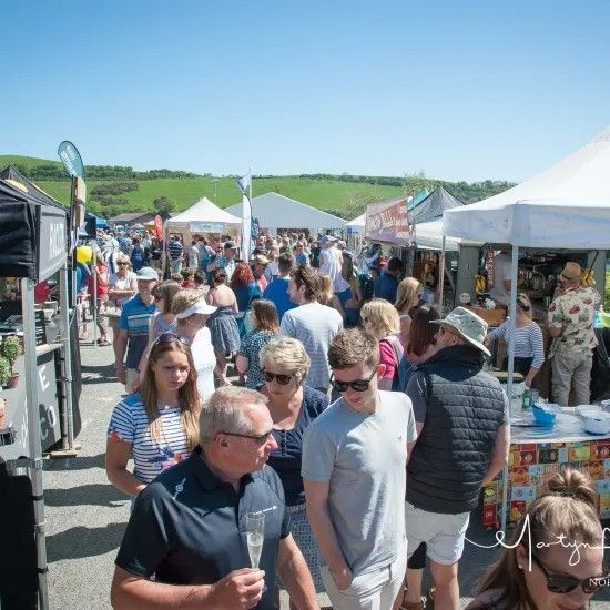 Crowded outdoor market on a sunny day. People browse vendors' tents, enjoying food, drinks, and the weather.