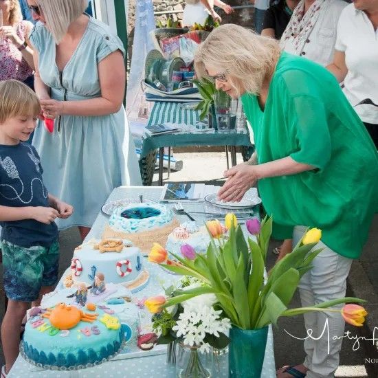 Woman in green examines cakes at an outdoor table; child looks on. Brightly colored cakes, flowers, and other people nearby.