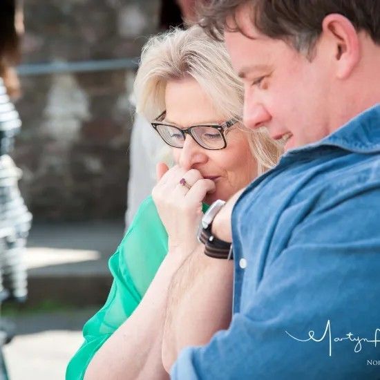 Woman in glasses and green top looks at watch with man in blue shirt. Outdoors.