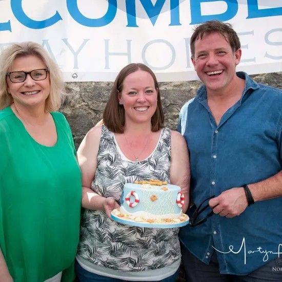 Three people smiling, holding a cake decorated with a beach theme, outdoors.