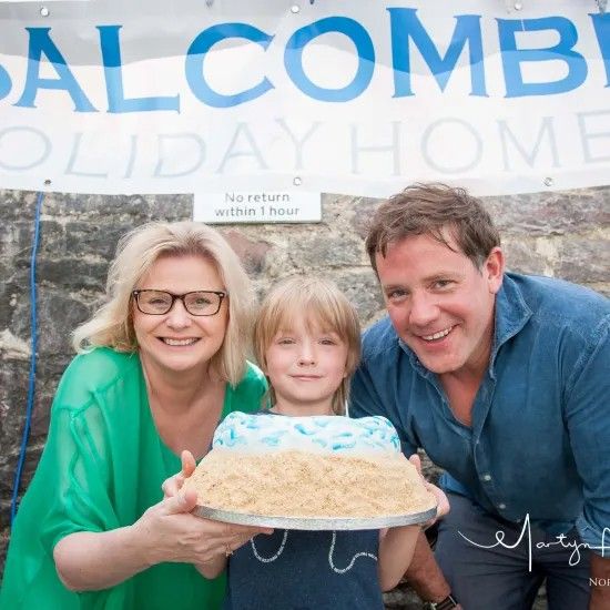 Family holding a cake. Banner reads 'Salcombe Holiday Homes.' Woman, boy, and man smile. Blue and tan cake.
