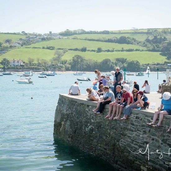 People sitting on a stone pier, overlooking a bay with boats and green hills on a sunny day.