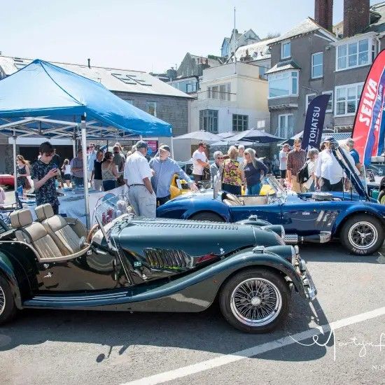 Classic cars on display at an outdoor event; spectators mill about. Blue tent, sunny day.