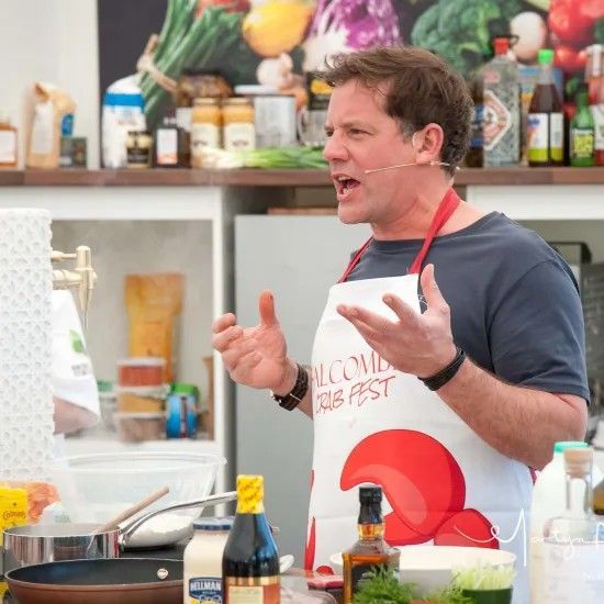 Man in apron speaking at a cooking demonstration, with food and ingredients visible.
