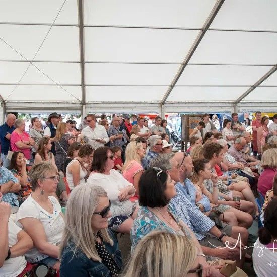 People seated in a tent listening to a speaker, with a crowd of people in the background.