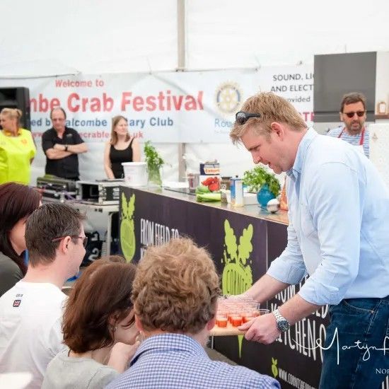 Chef at cooking demo: slicing tomatoes at the Salcombe Crab Festival. Attendees watch, stage visible, tents, banner.