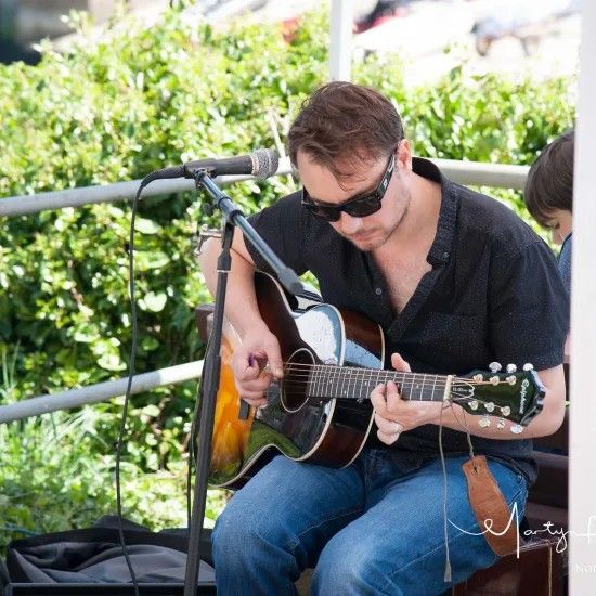 Man playing acoustic guitar, wearing sunglasses and black shirt, performing outdoors.