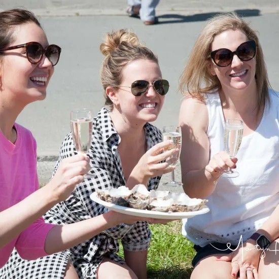 Three women in sunglasses laugh while holding champagne glasses near a plate of oysters. Outdoors.