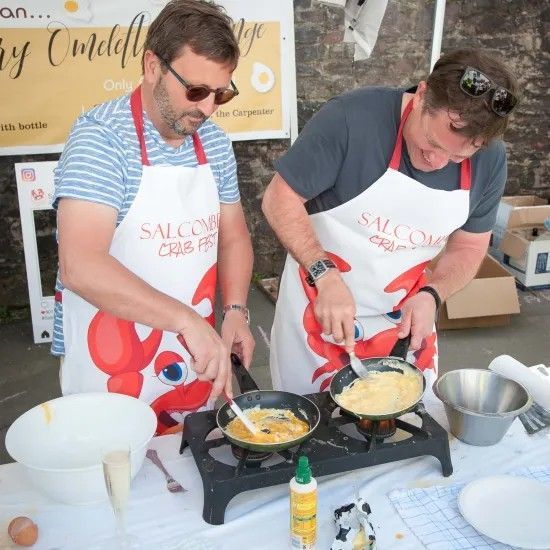 Two men cooking scrambled eggs at an outdoor event, wearing aprons, with pans on a portable stove.