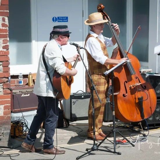 Two musicians playing instruments outside. One plays guitar, the other a double bass. Sunny day.