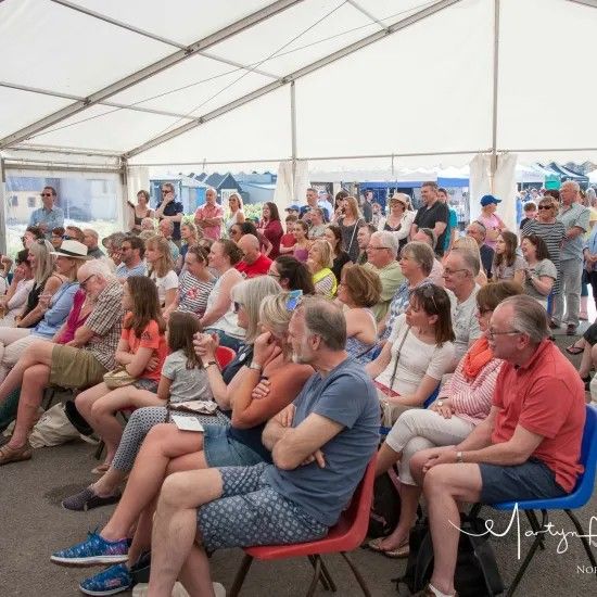 People seated under a tent listening to an event, some are smiling.