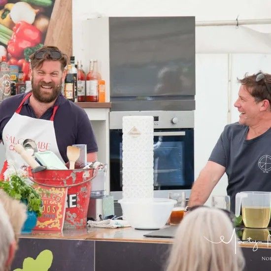 Two men smiling, cooking at a food stall, one wearing an apron, in front of an audience.