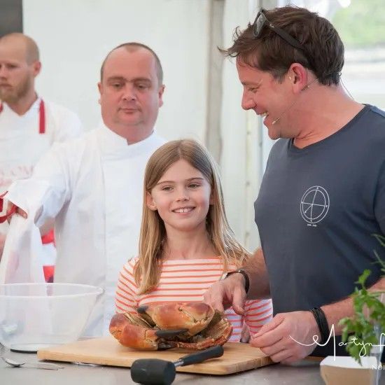 Chef, girl, and man at a kitchen counter with food. Girl smiles, man points, chef looks on.