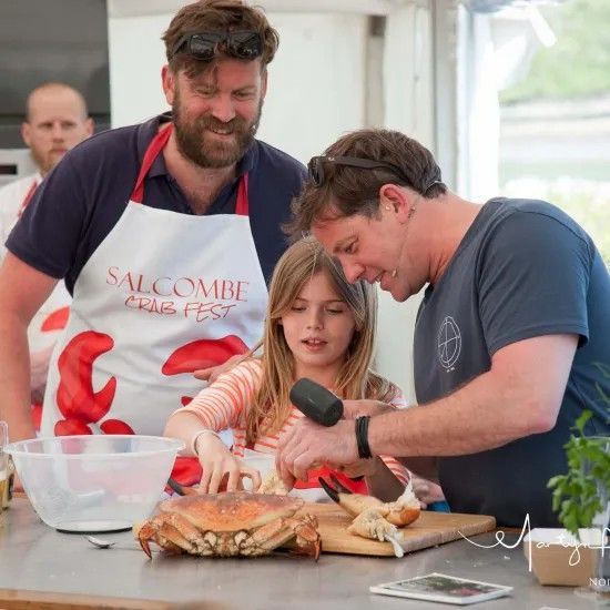 People cracking crab at a cooking event; a girl, two men. One man uses a mallet. A 