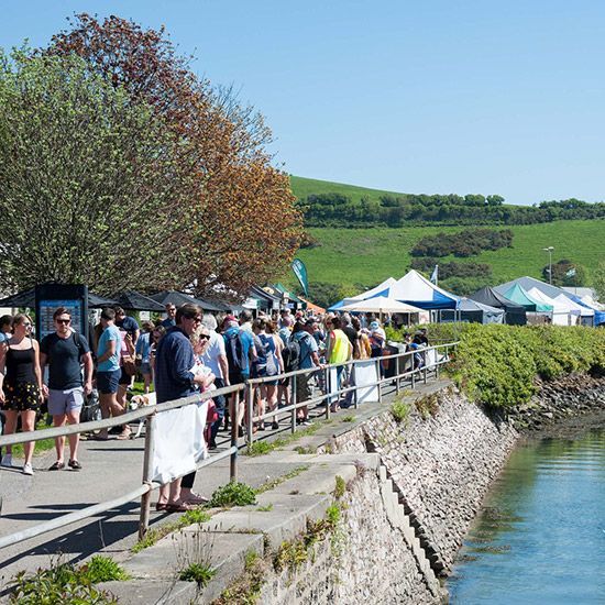 People walking along a waterfront promenade, blue sky, tents set up, green hills in the background.