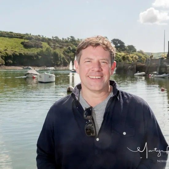 Man smiles in front of a harbor with boats on a sunny day.