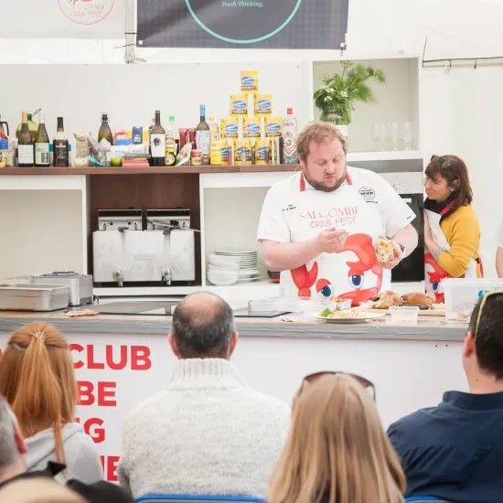 Chef preparing food at a demonstration; people watch. Counter with bottles and products.