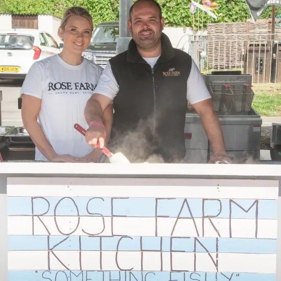 Two people stand behind a food stall. Woman in white shirt, man in black vest, preparing food. Stall sign: 